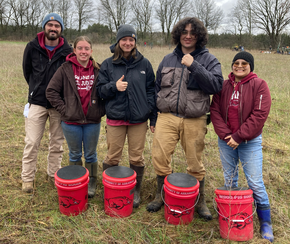 Five people standing in a field in front of four red razorback buckets.