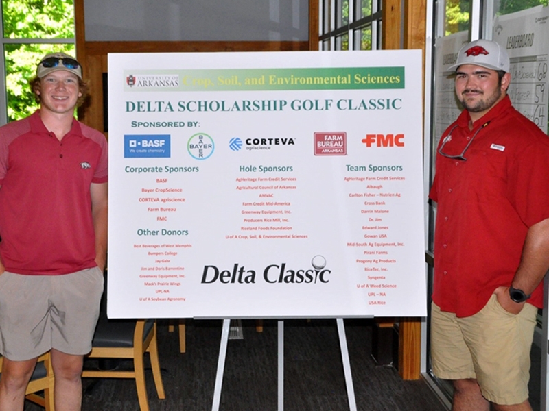 Two men in red polo shirts and khaki shorts standing on opposite sides of the Delta Scholarship Golf Classic sponsors poster