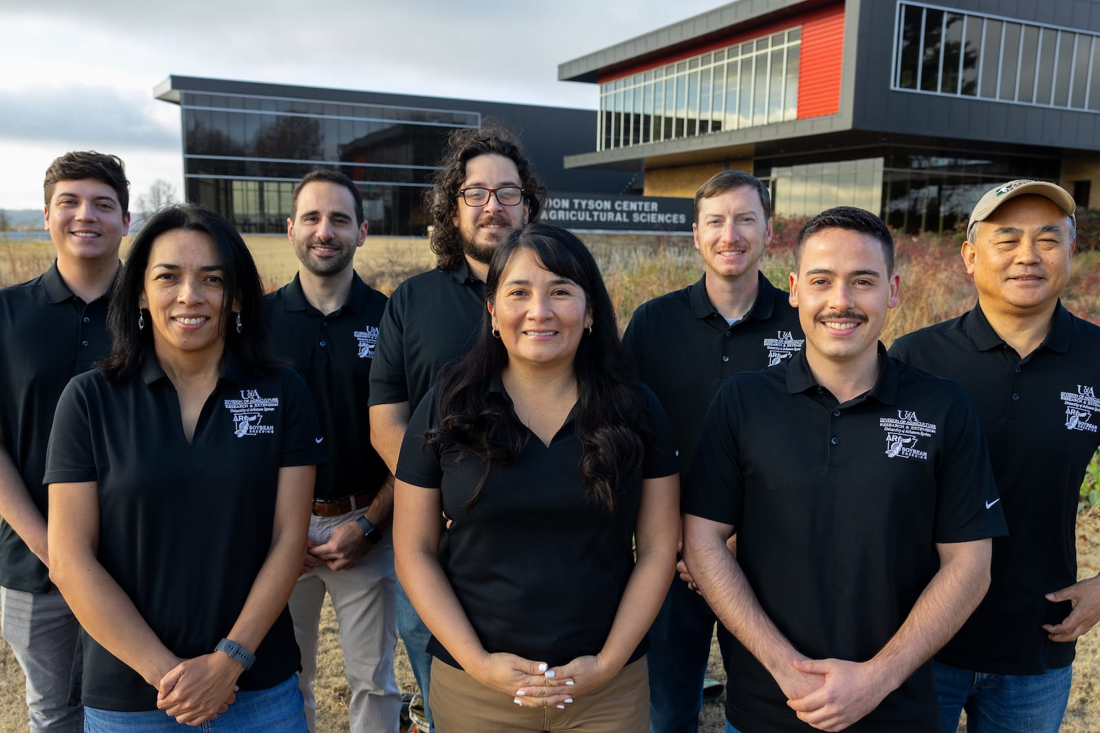 Two rows of people (5 men in the back, 2 women and 1 man in the front) all in black polo shirts standing in front of Don Tyson Center for Agricultural Sciences