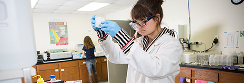 Female student wearing lab coat and goggles measuring liquid